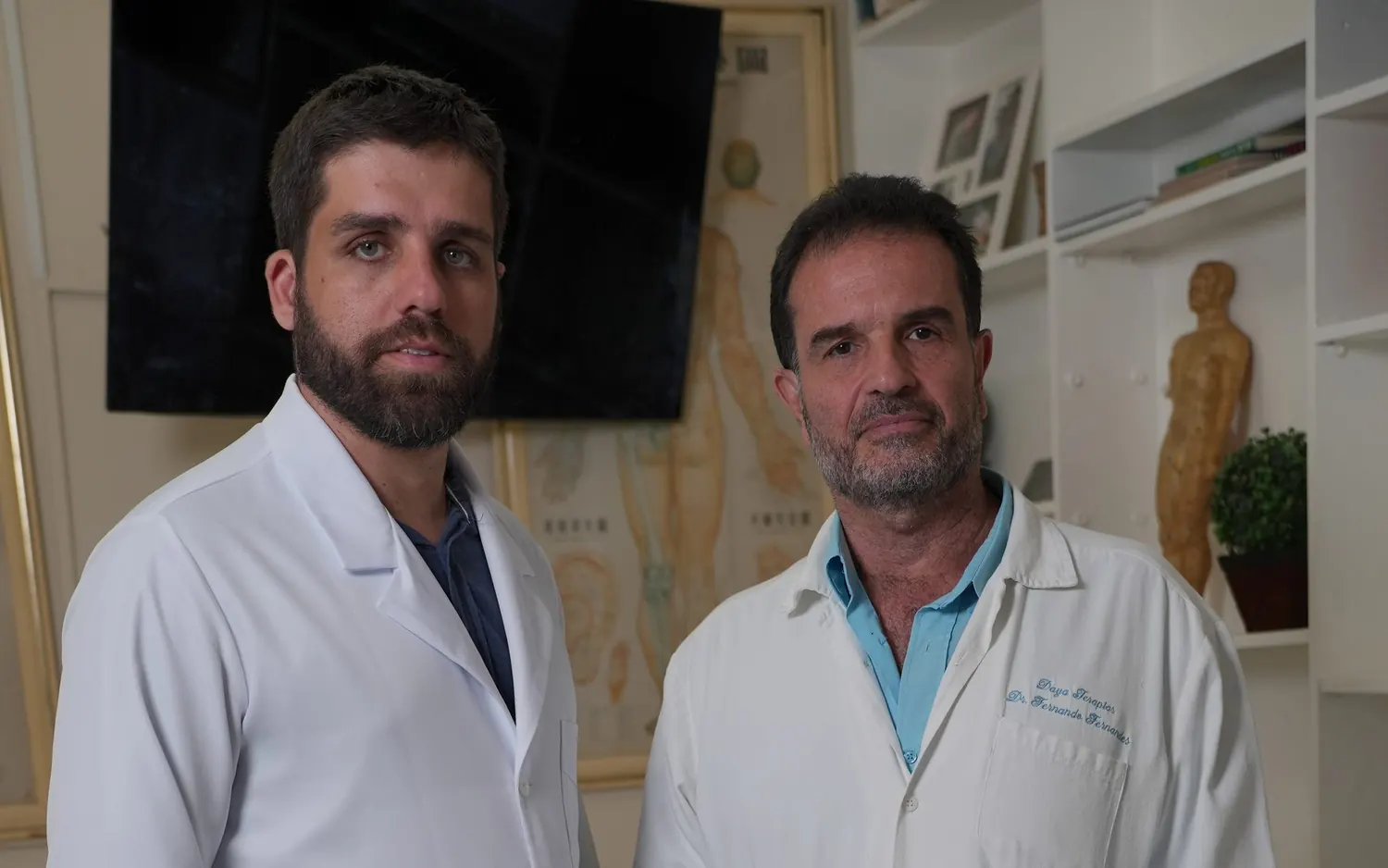Os dois doutores de jaleco no consultório, com mapas de acupuntura na parede