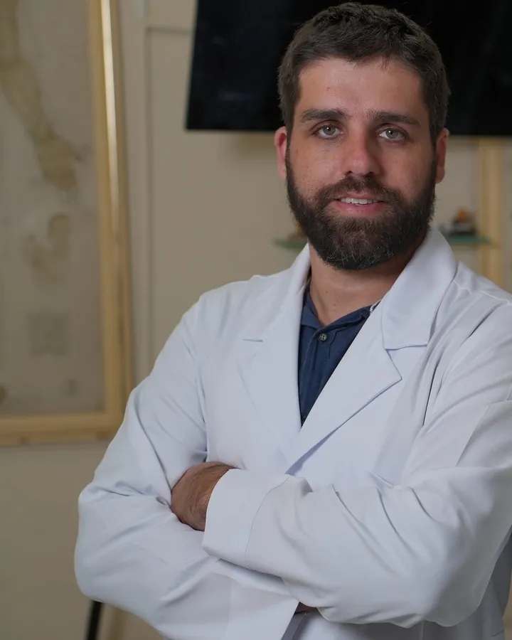 Dr. Rodrigo Damásio de jaleco, braços cruzados, no consultório